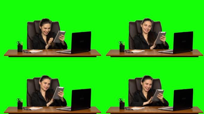 工人女孩坐在工作场所，靠在皮椅上，在平板电脑上交流，撒娇地微笑。绿屏