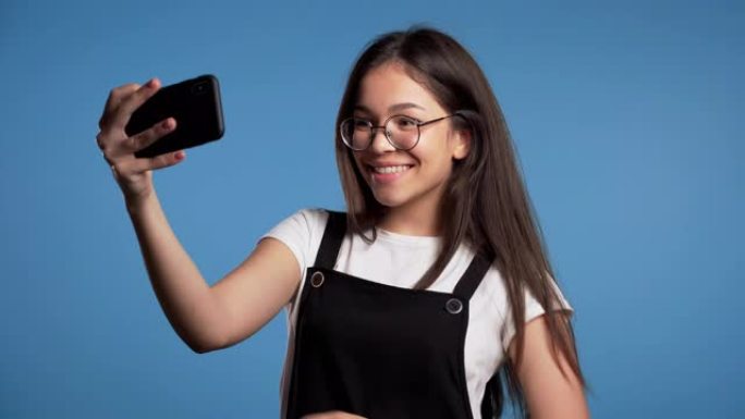 戴着眼镜微笑的快乐女孩在蓝色背景上的智能手机上自拍。技术、移动设备、社交网络概念。