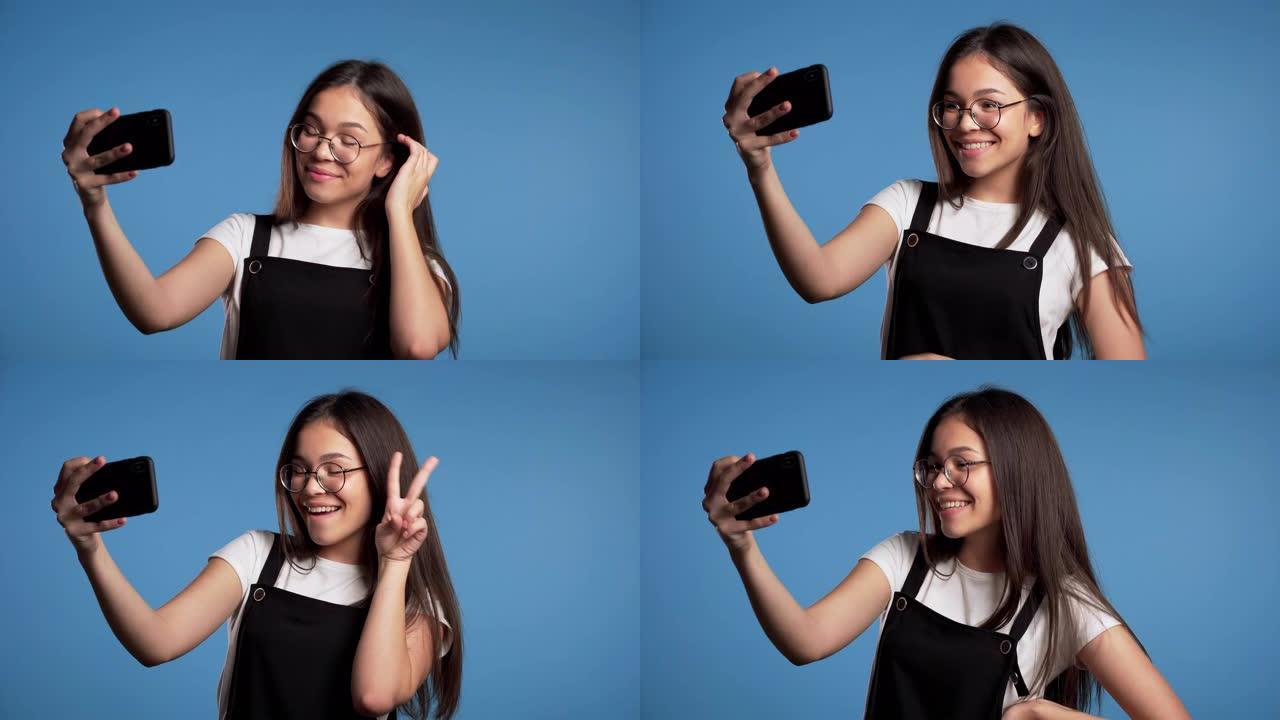 戴着眼镜微笑的快乐女孩在蓝色背景上的智能手机上自拍。技术、移动设备、社交网络概念。