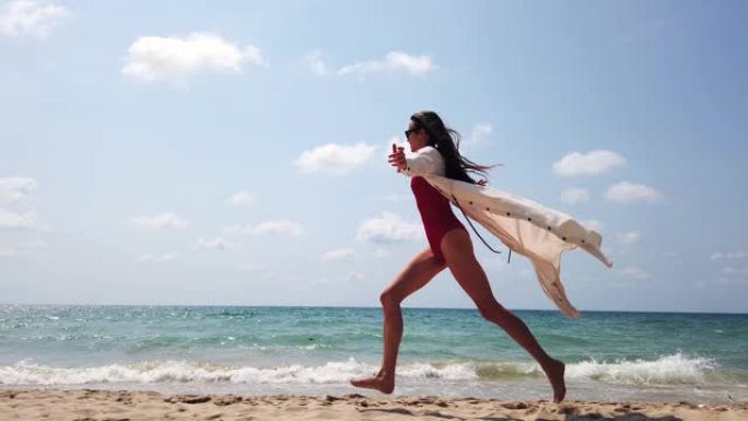 穿着红色泳衣，白色长裙，戴着太阳眼镜的年轻女士在海边的沙滩海岸线上奔跑，波涛汹涌。透过棕榈叶。暑假快