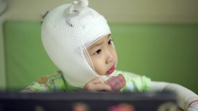 亚洲小女孩在餐桌上吃零食生病了。亚洲女婴头部和手臂包扎生病。手持镜头，真实生活