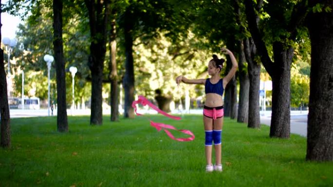 在一个阳光明媚的夏日，一个穿着运动服装的白人女孩站在芭蕾舞姿势，在城市公园的草地上扭动体操丝带。慢动