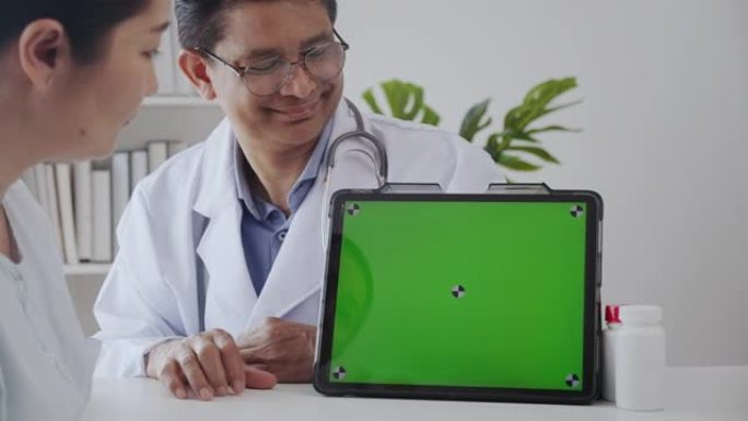 高级医生向患者解释在诊所办公室使用绿屏数字平板电脑并微笑。