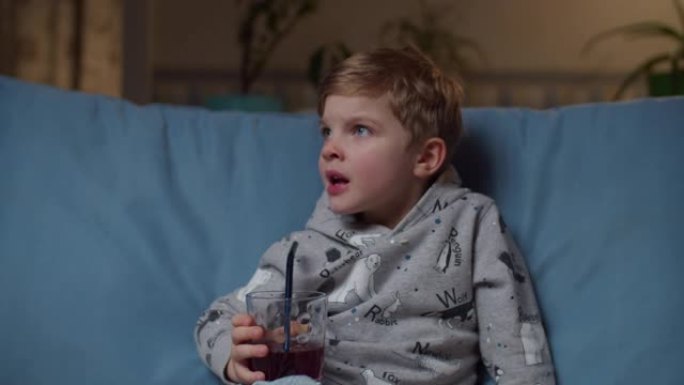晚上坐在舒适的沙发上喝果汁的学龄前男孩的特写镜头。孩子在家沙发上用非塑料吸管喝蓝莓汁。