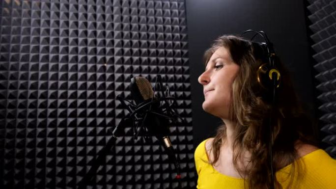 戴着耳机的女人对着黑色背景的麦克风唱歌。穿着黄色衣服的专业女歌手。在音乐工作室录制一首歌。
