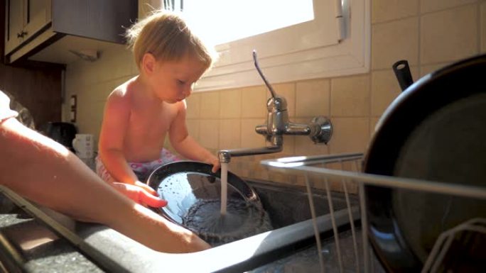 妈妈洗碗时，孩子坐在水槽里。母女概念。厨房职责。家务和孩子。正在工作的母亲和蹒跚学步的孩子。母亲的手