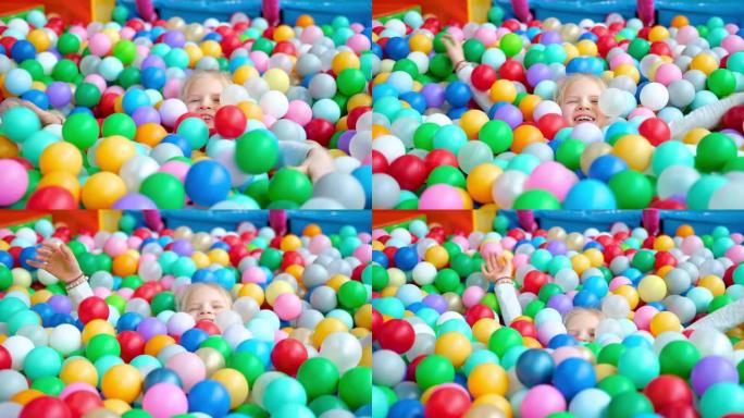 可爱的金发小女孩躺在游戏中心大干戏水池里的多色塑料球上。对着镜头微笑。肖像特写。在游戏室玩得开心。休
