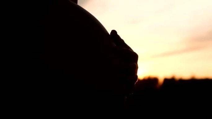 一名孕妇在日落时用手抚摸腹部的特写剪影。