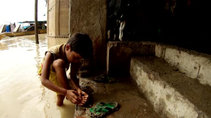 印度小男孩在淹没的恒河的肮脏水中洗脚，在牛屎瓦拉纳西的码头拖鞋上的牛船蹄子