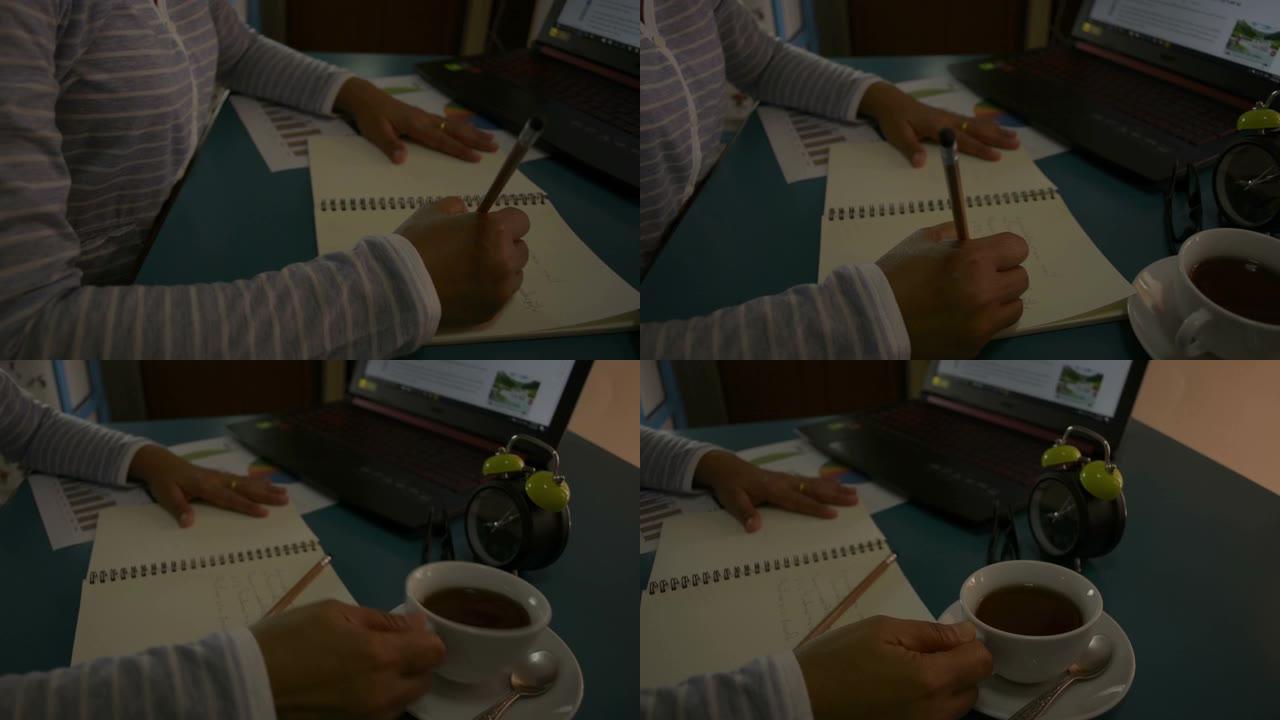 用笔记本电脑在桌子上工作的女商人。女人的手在记事本上写字，在家喝一杯咖啡。