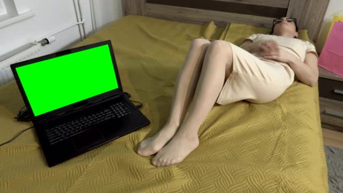 女人下班后回家脱下高跟鞋，在绿色屏幕笔记本电脑附近的床上放松
