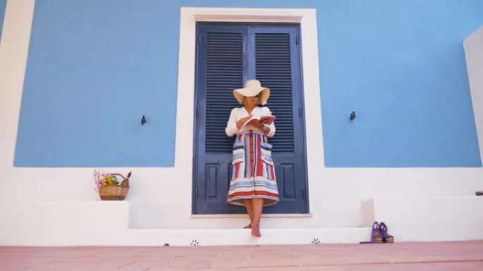 年轻快乐的女人站在蓝色房子入口露台的台阶上看书。时尚白衬衫，大帽子，彩色裙子和太阳镜。意大利蓬扎岛。