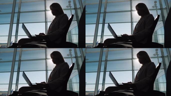一位带着笔记本电脑的女商人的剪影，期待着她的飞行。坐在机场航站楼的大窗户旁