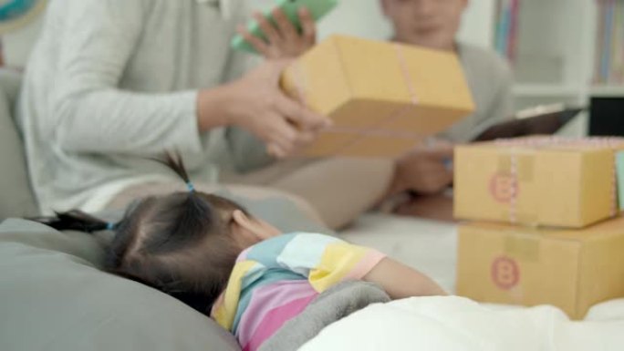 4k慢动作，年轻的亚洲家庭通过平板电脑和包装产品检查客户的订单，而女婴则睡在床上。商务电子商务在家概
