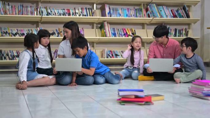一群亚洲小孩从老师那里学习使用笔记本电脑，坐在学校图书馆的地板上，背景是书架