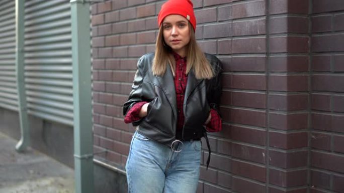 美丽的欧洲女孩穿着黑色皮夹克和牛仔裤和红色帽子站在后院口袋里靠着红色砖墙