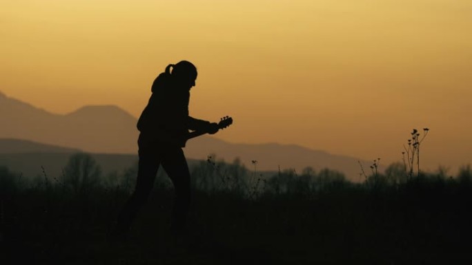 一个年轻的女人带着吉他在日落时在大自然中演奏音乐。轮廓。