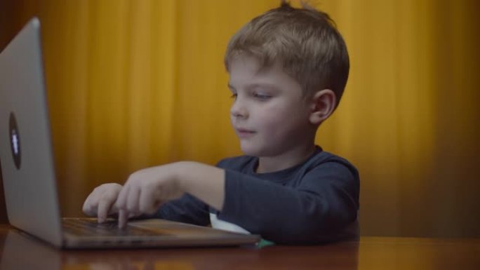 金发碧眼的学龄前男孩在笔记本电脑上打字，微笑着看着电脑屏幕。孩子坐在桌旁在家在线学习。