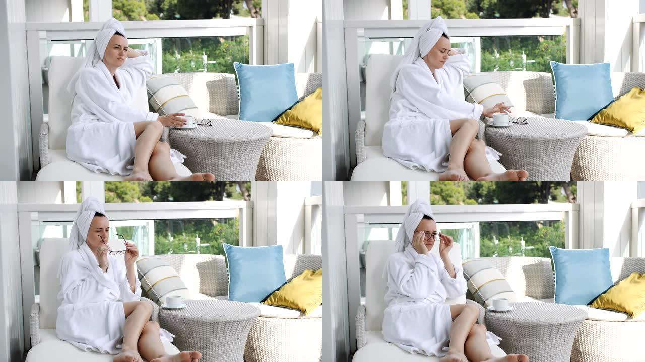穿着浴袍的美女躺在露台上的日光浴躺椅上喝咖啡，洗个热水澡后在新鲜空气中放松。