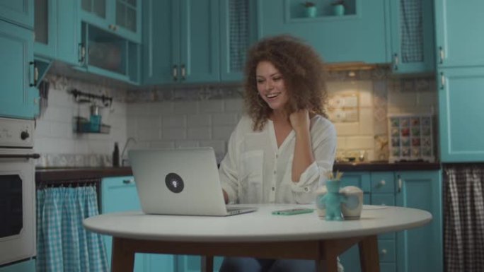 年轻的20多岁的女人在厨房的笔记本电脑上进行视频通话。女性在笔记本电脑屏幕上动情地说话，双手示意，坐