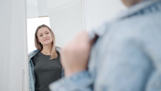 白色房间里一位年轻迷人的千禧一代女性试穿并选择衣服。照镜子，比较服装。房子的衣柜间或店内的试衣间。