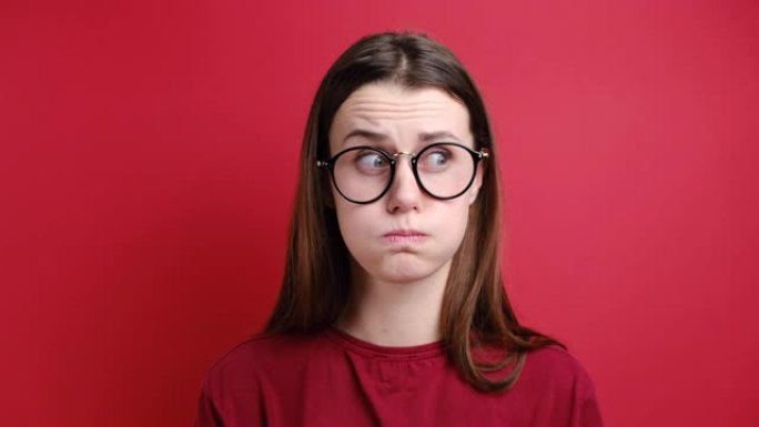 戴眼镜的怨恨女孩在嘲笑表现出负面情绪和怨恨的人时吹着脸颊，穿着t恤，站在红色背景上