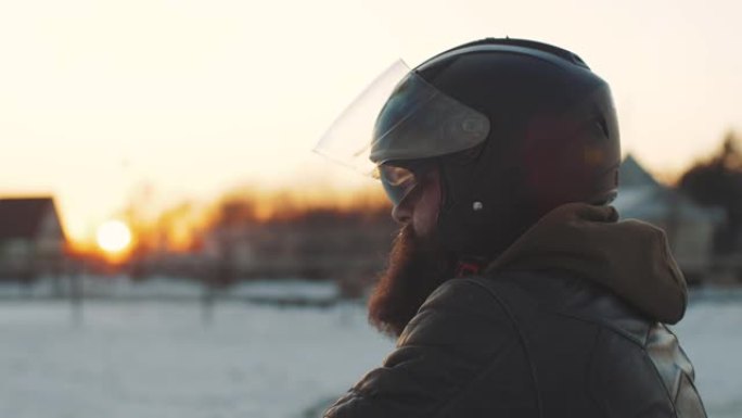 日落时在雪场上戴着黑色头盔的男子摩托车手的肖像。男子在寒冷的冬天骑摩托车