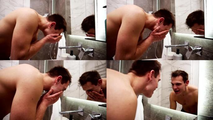 赤裸上身的帅哥在水龙头的冷水下在水槽里洗脸。肌肉发达的人做早上例行清洁程序。看着镜子的倒影和微笑。慢
