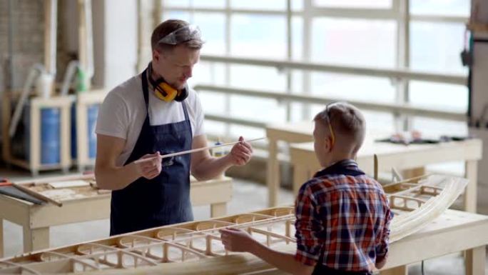 中年木匠的中等跟踪镜头教小儿子如何在木工车间的工作台上建造木船