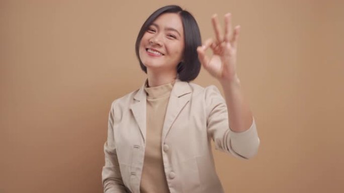亚洲女商人快乐的微笑展示她的手在米色背景上孤立的OK手语。4k视频