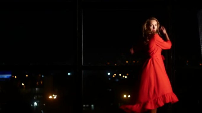 一个穿着优雅的红色连衣裙的女孩在一个傍晚城市的大窗户的背景下跳舞。一个人在家，一个人相爱