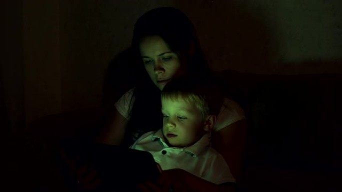 母亲和儿子在黑暗中在家中的平板电脑的肖像。