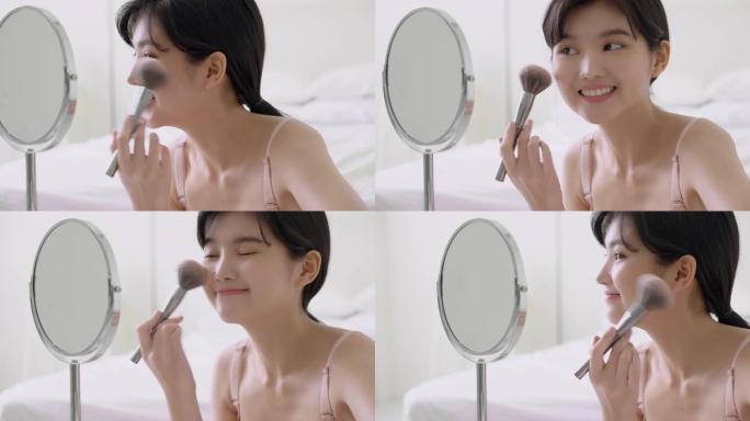 美丽的年轻亚洲女人快乐和微笑与面镜在房间里用脸颊刷化妆，美丽的女孩拿着刷子化妆在面部，皮肤护理和美容