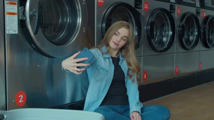 快乐的年轻女子在自助洗衣店的智能手机上自拍。自助公共洗衣房。