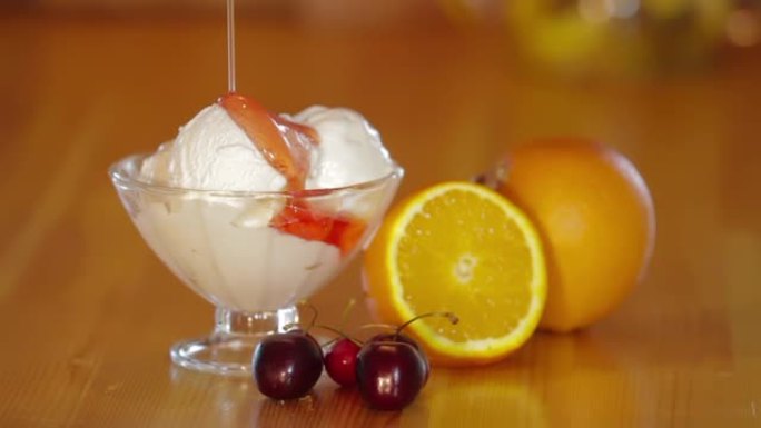 玻璃碗里的冰淇淋和木桌上的橙子和樱桃。冰淇淋冰淇淋的制备。甜冷甜点。五颜六色的冰淇淋球倒入红色果酱。