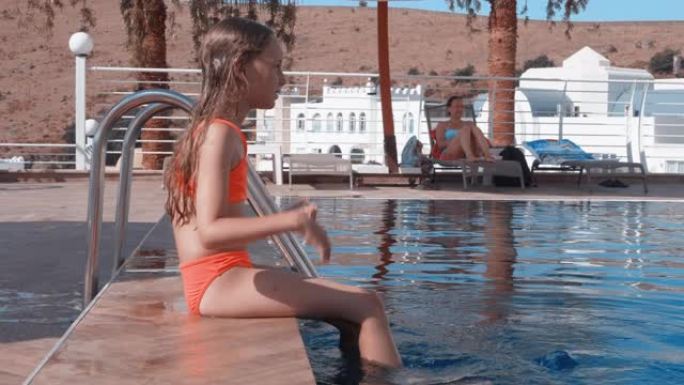 在度假酒店，十几岁的女孩坐在泳池边缘，将双腿浸入水中。年轻女孩在夏季酒店用脚在游泳池里泼水。旅游女孩