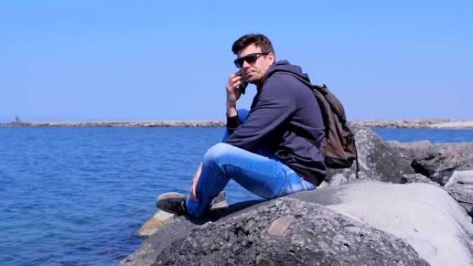 男人旅行者打电话给智能手机坐在石头上等待海上背景的答案。