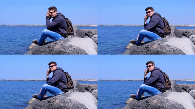 男人旅行者打电话给智能手机坐在石头上等待海上背景的答案。