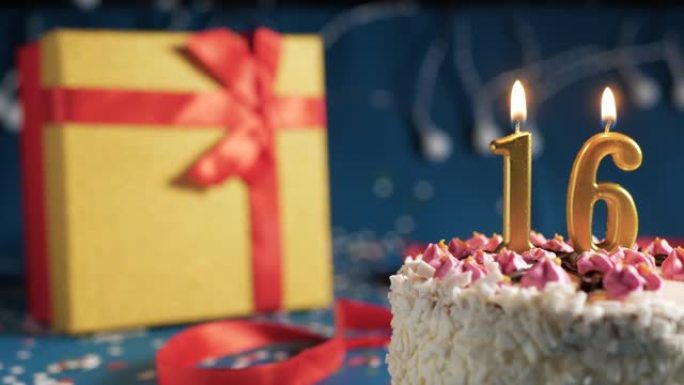 白色生日蛋糕编号16点灯燃烧的金色蜡烛，蓝色背景带灯和用红丝带绑起来的礼物黄色盒子。特写