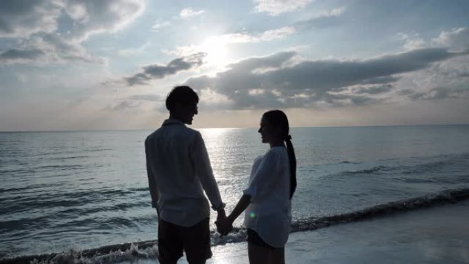 后视图快乐的亚洲年轻夫妇在海滩日落背景上手牵手并进行眼神交流。假日和旅游概念。慢动作