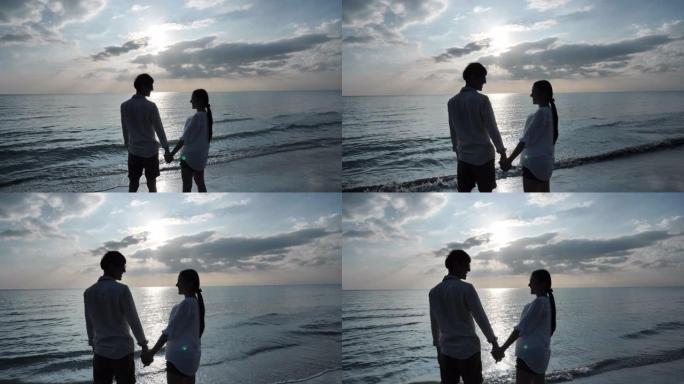 后视图快乐的亚洲年轻夫妇在海滩日落背景上手牵手并进行眼神交流。假日和旅游概念。慢动作