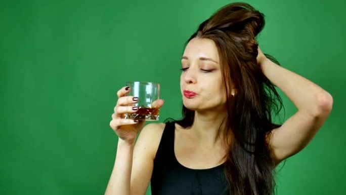 年轻的黑发悲伤的女人，长长的黑发正在工作室里从绿色背景的玻璃杯里喝干邑白兰地