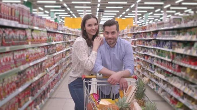 迷人的快乐年轻千禧一代家庭夫妇用购物车在超市购物，看着相机微笑，可爱的夫妻买家顾客满意杂货店概念