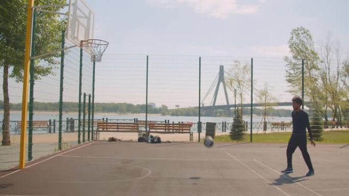 运动型迷人的非洲裔美国男子篮球运动员的肖像把球扔进篮筐，在城市的户外球场上不开心，背景是布里奇