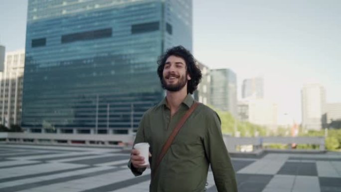 微笑的年轻男性专业人士在市中心散步时喝咖啡，背景是现代企业建筑