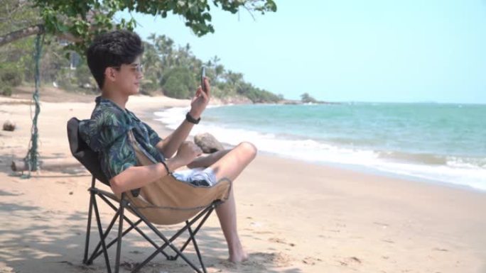 4k年轻的亚洲男子坐在热带岛屿海滩的沙滩椅上，使用智能手机与视频通话移动应用程序与女友交谈。具有户外