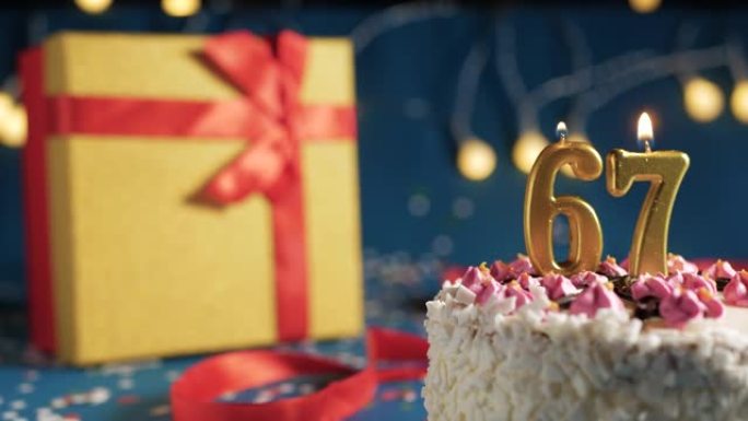 白色生日蛋糕67号点火器燃烧的金色蜡烛，带灯的蓝色背景和用红丝带捆绑的礼物黄色盒子。特写