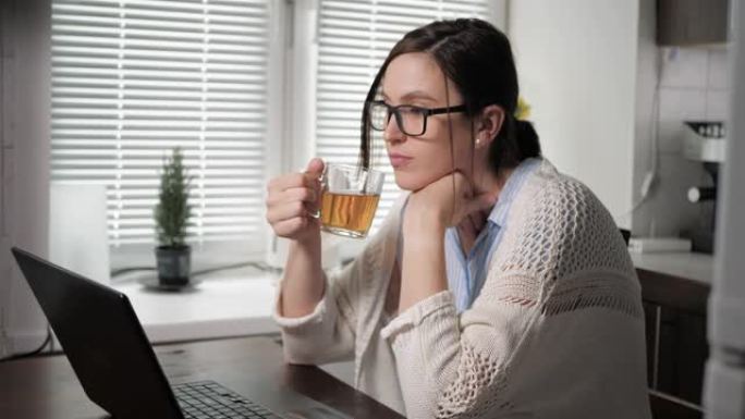 戴着眼镜和针织毛衣的迷人白人女孩在公寓的厨房里，坐在桌子旁喝茶，看着笔记本电脑屏幕上的东西。自由职业