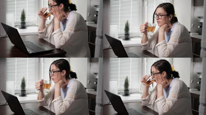 戴着眼镜和针织毛衣的迷人白人女孩在公寓的厨房里，坐在桌子旁喝茶，看着笔记本电脑屏幕上的东西。自由职业