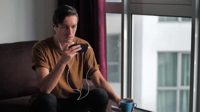 戴着耳机的微笑男人坐在家里的沙发上喝美味的咖啡，享受着智能手机在社交网络中观看媒体的乐趣。